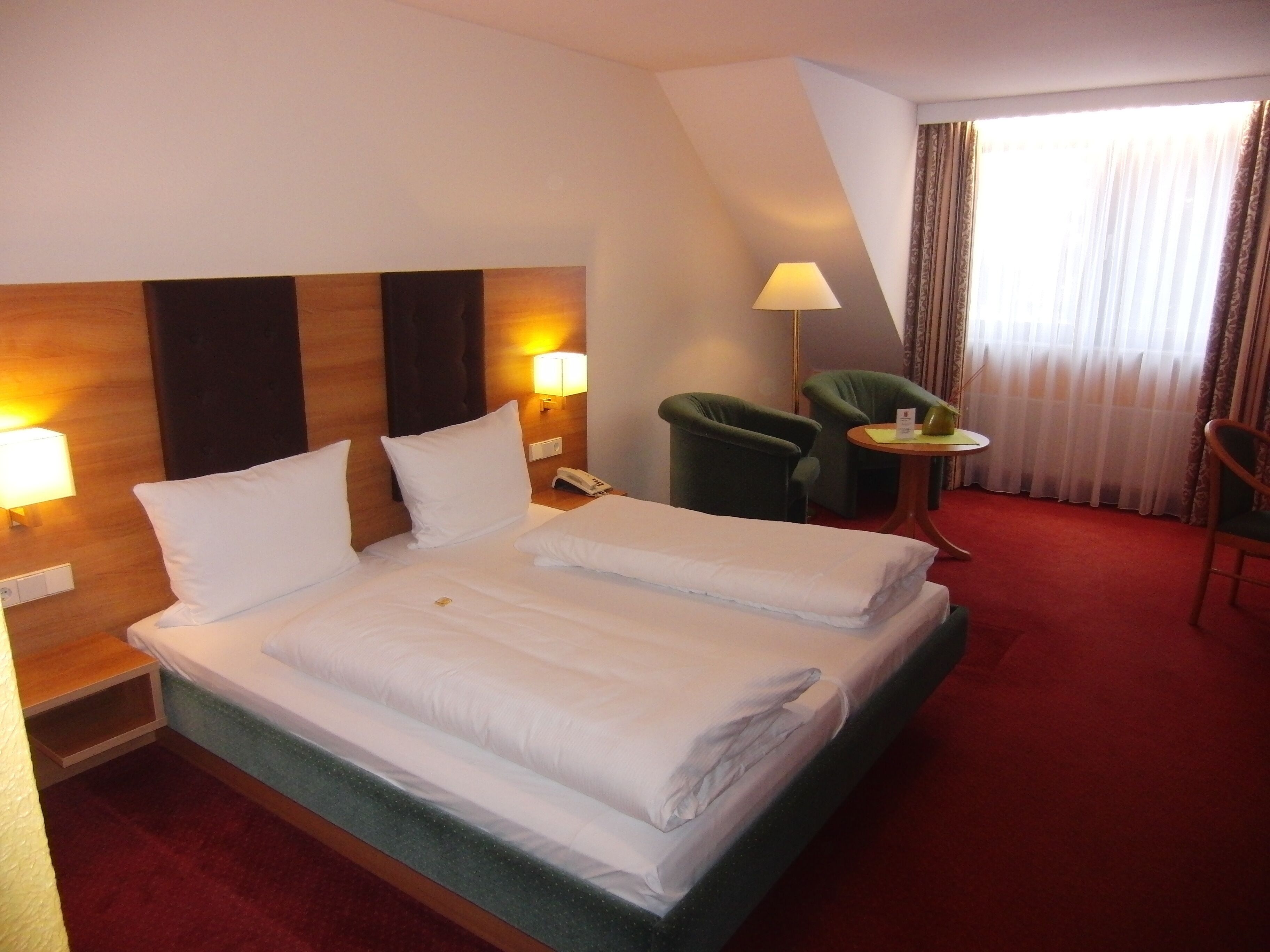 A hotel room with a neatly made bed, two bedside lamps, a telephone, a small table with a lamp, two chairs, and a large window with curtains.