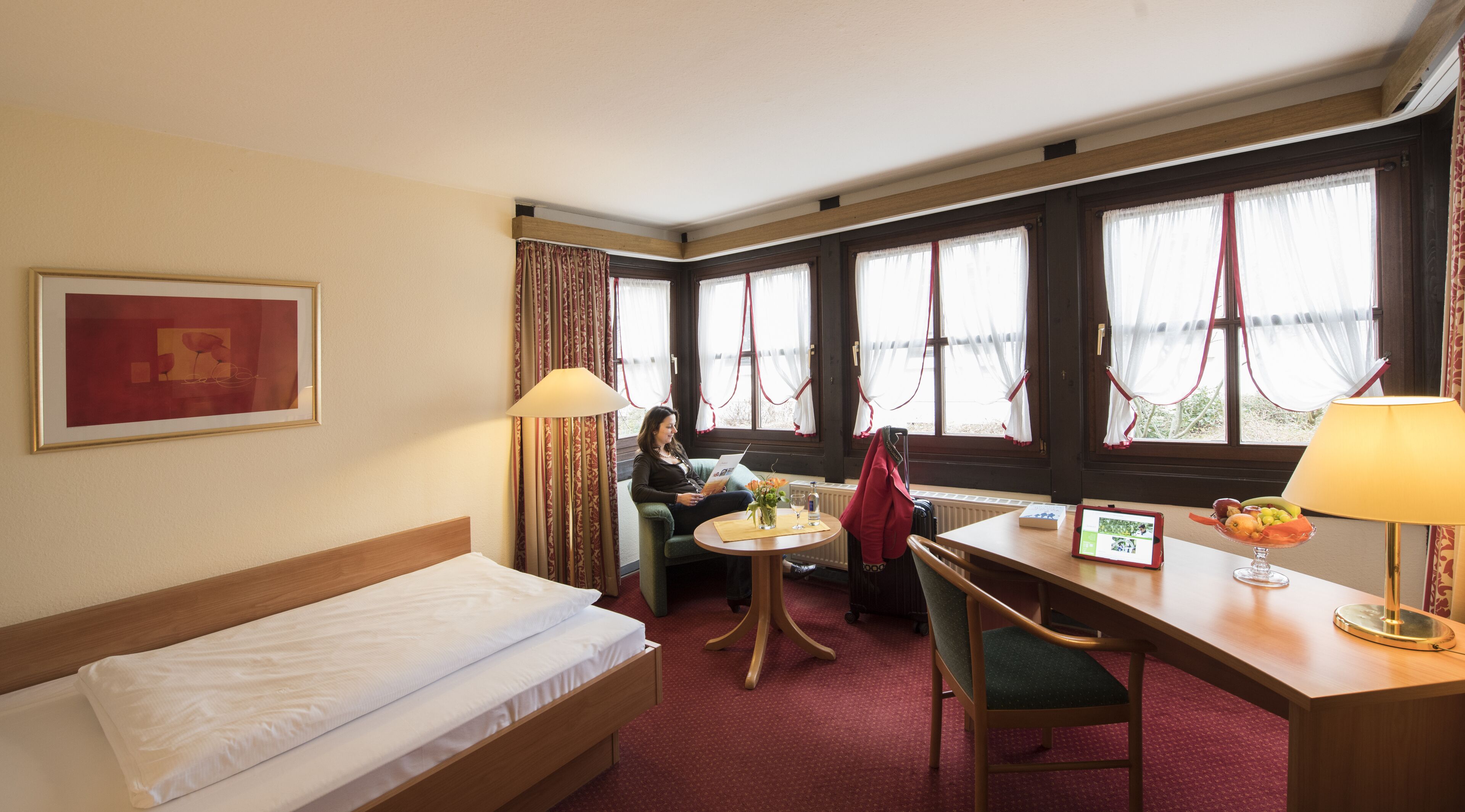 Une chambre d’hôtel comprenant un lit, un bureau avec une chaise, une table avec un vase et une tablette, ainsi que de grandes fenêtres avec des rideaux.