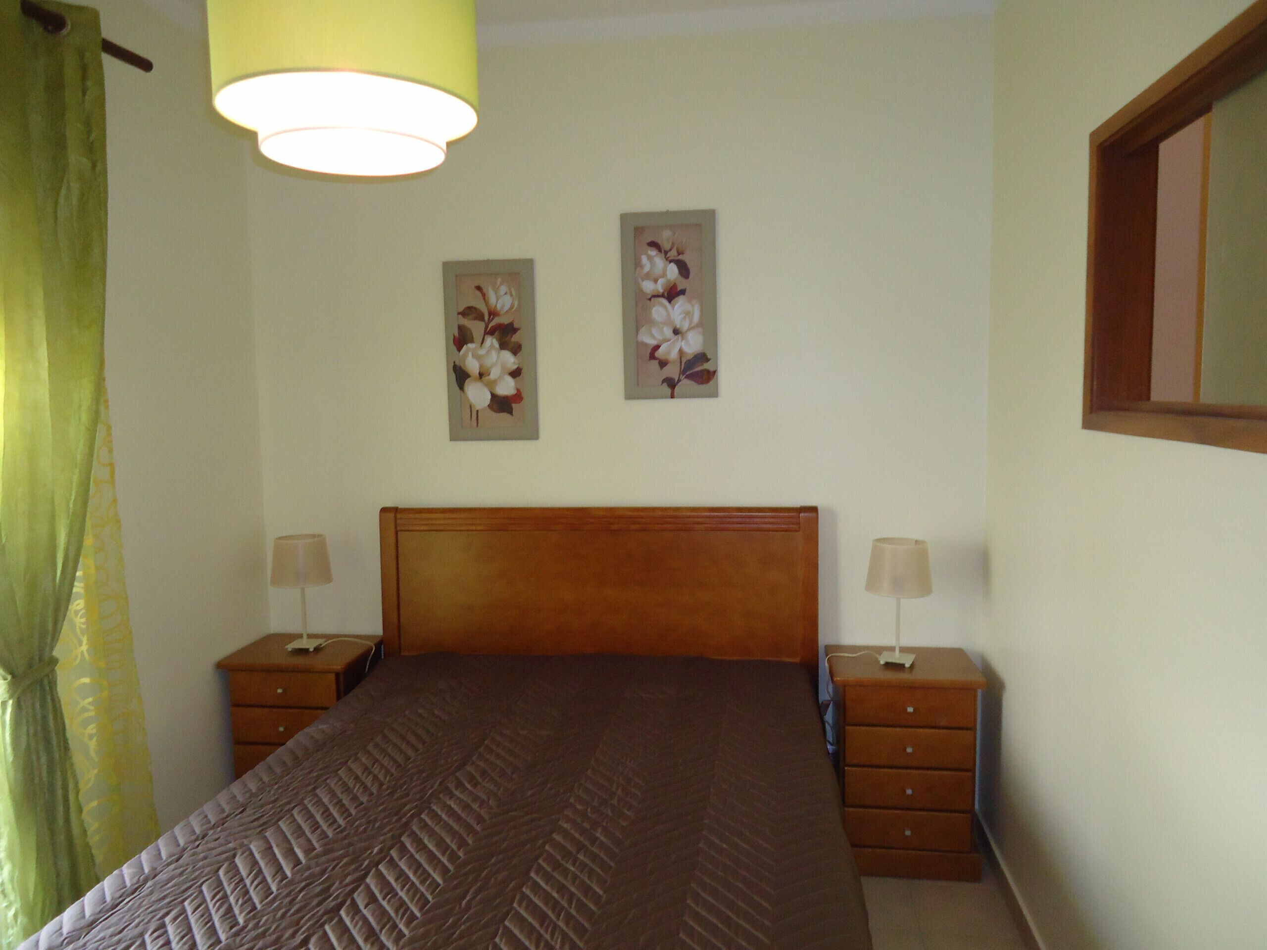 A bedroom with a wooden headboard, two bedside tables with lamps, a bed with a patterned bedspread, and two wall-mounted floral paintings.