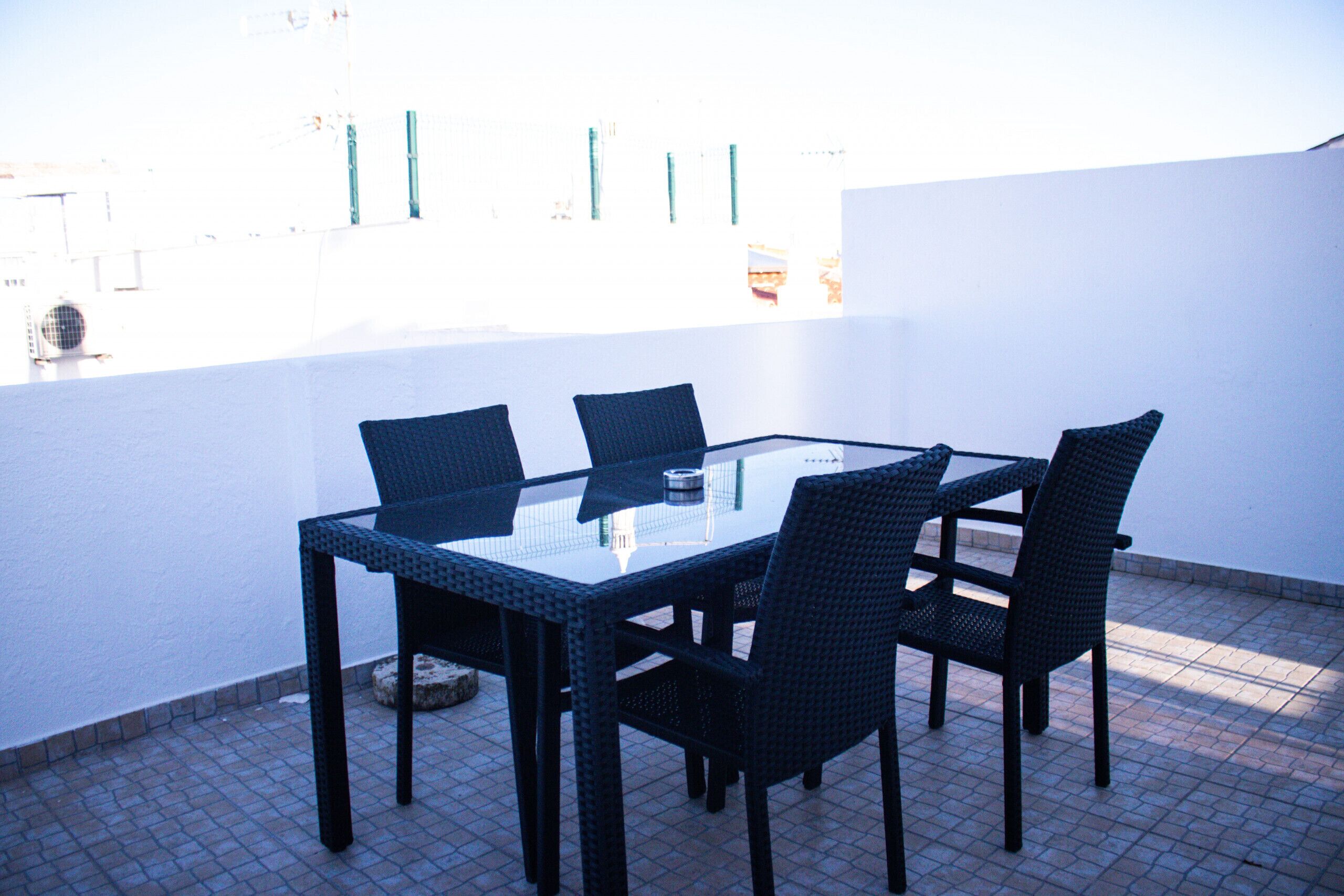 A glass-top table with four chairs, set on a tiled patio with a white wall in the background.