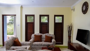 A living room with a sofa, a coffee table, and a television. There are three windows with wooden shutters. - Villa Cannelles (Micoud)