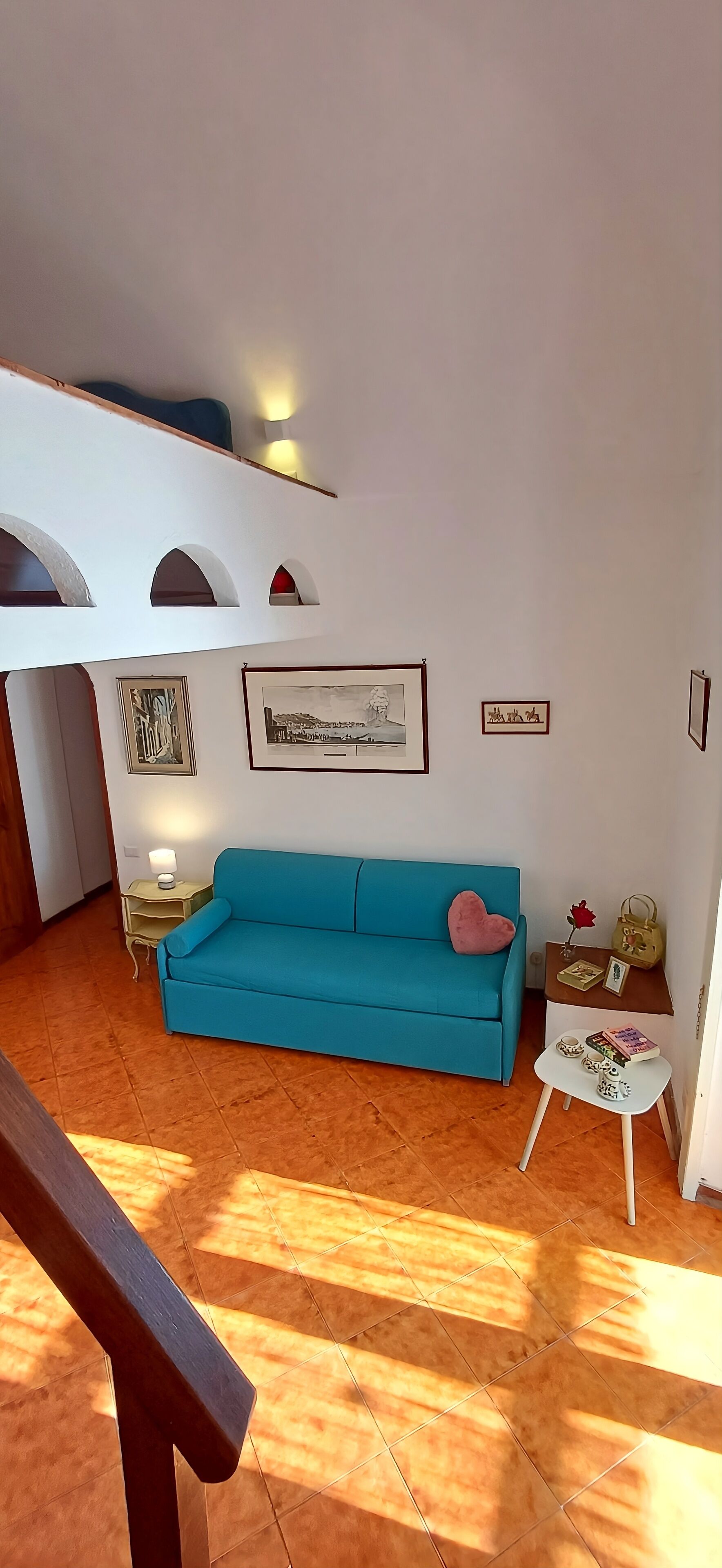 A room with a blue sofa, a small white table with books and a decorative item, a wall with framed pictures, and a staircase with a wooden railing.