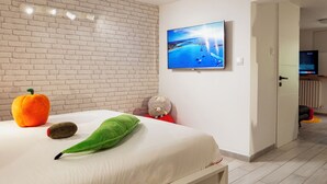 Una camera da letto con parete in mattoni, un letto con cuscini, una televisione che mostra una spiaggia e una porta che dà su un'altra stanza.