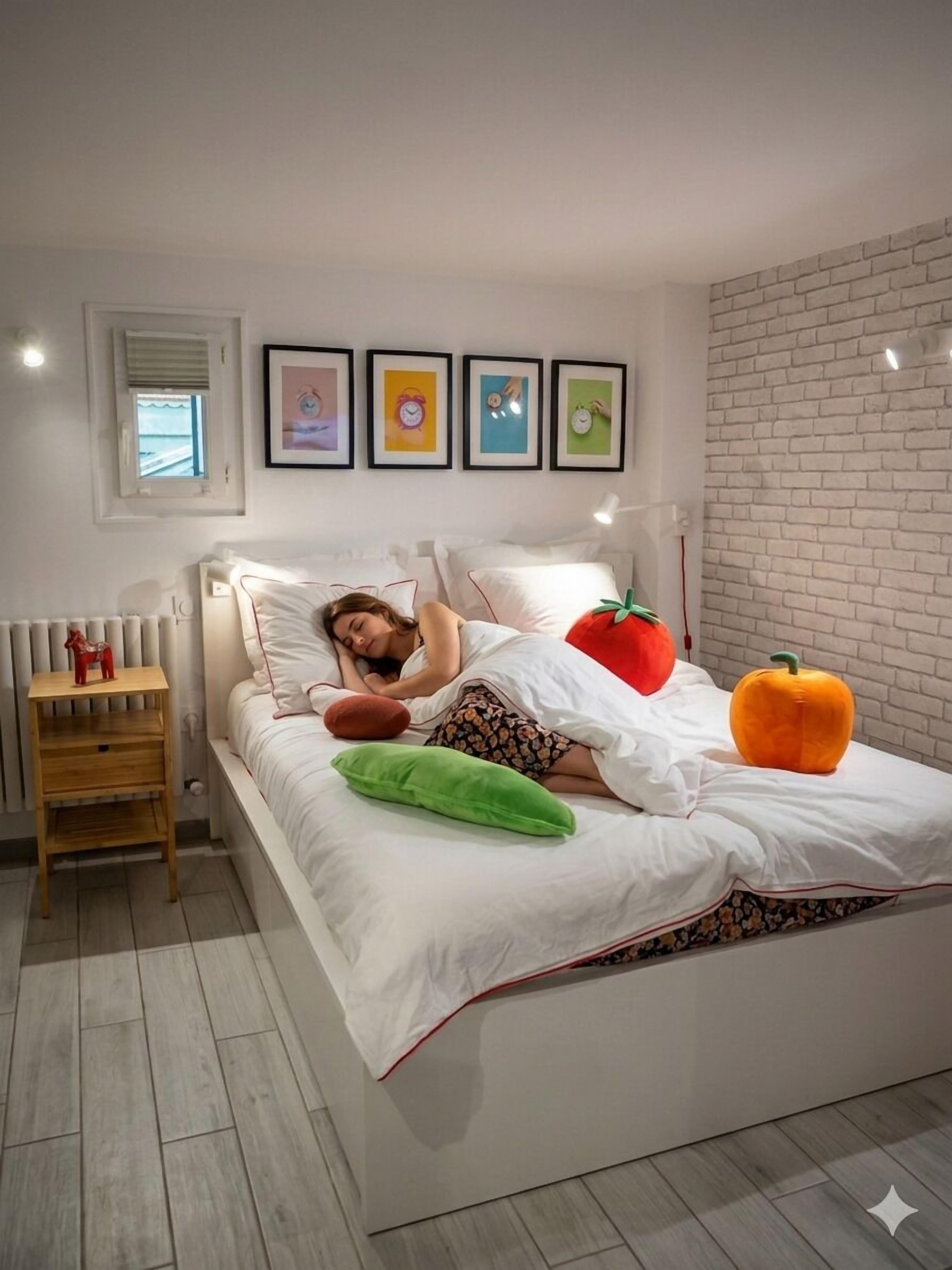 Un dormitorio con cama, almohadas, mesita de noche, radiador y cuadros en la pared.
