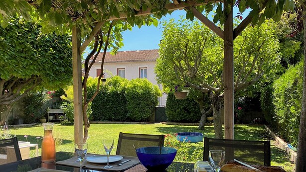 Un tavolo da pranzo all'aperto apparecchiato con piatti, posate e una ciotola blu, circondato da vegetazione lussureggiante e un edificio sullo sfondo.