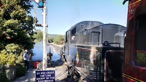 A vintage steam train at a railway crossing with a 'DO NOT CROSS THE LINE EXCEPT BY THE FOOTBRIDGE' sign.
