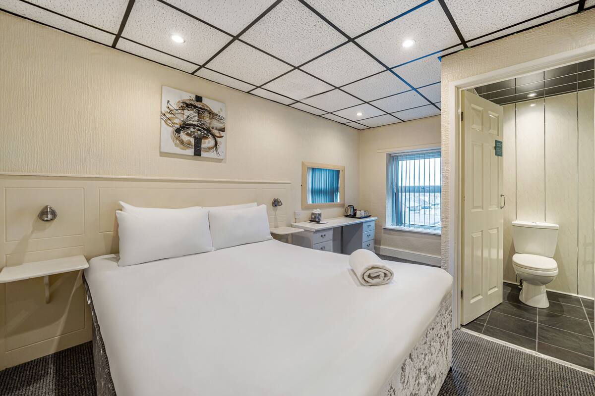 A hotel room with a large bed, a bedside table, a mirror, a small desk, and a toilet.