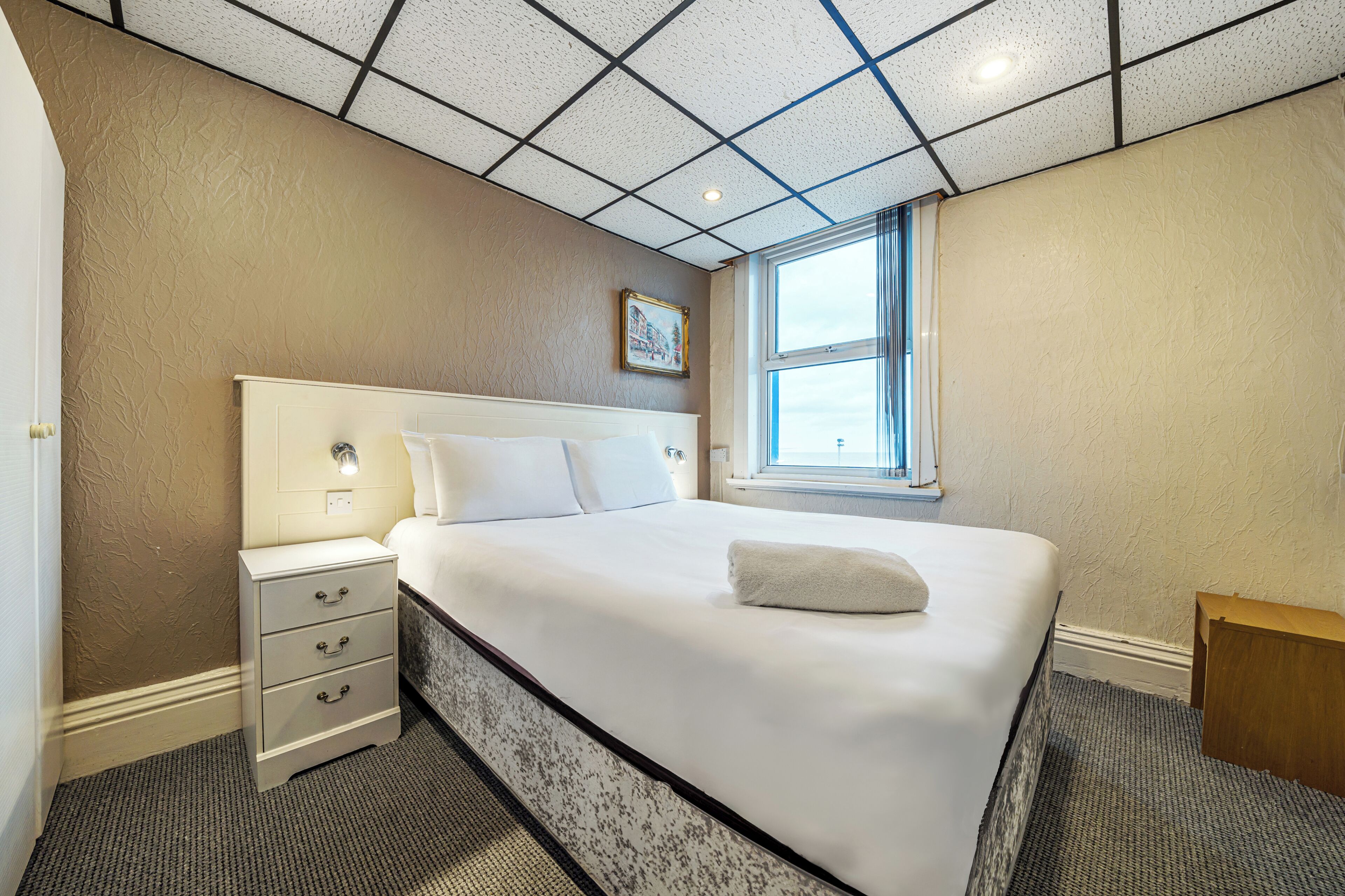 A neatly arranged bedroom with a large bed, a bedside table, and a window providing natural light.