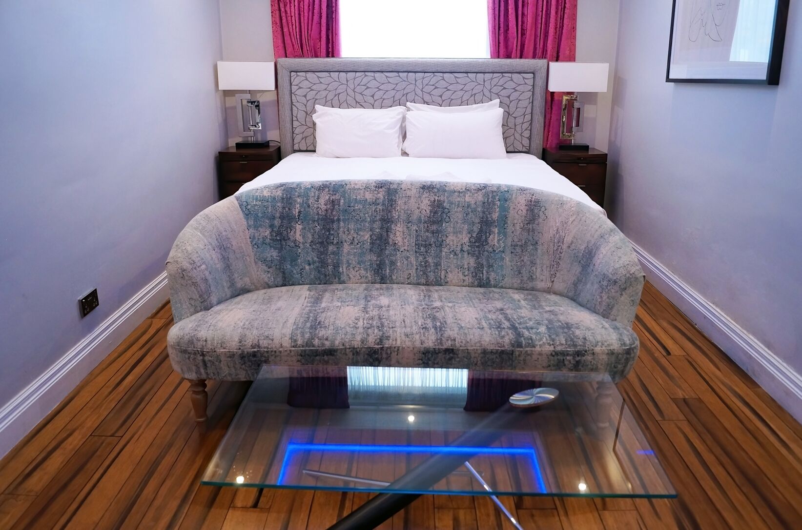 Un dormitorio moderno con una cama grande, un sofá, una mesa de centro de cristal con iluminación azul, suelo de madera y cortinas rosas.