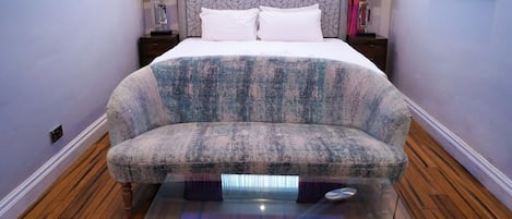Un dormitorio moderno con una cama grande, un sofá, una mesa de centro de cristal con iluminación azul, suelo de madera y cortinas rosas.