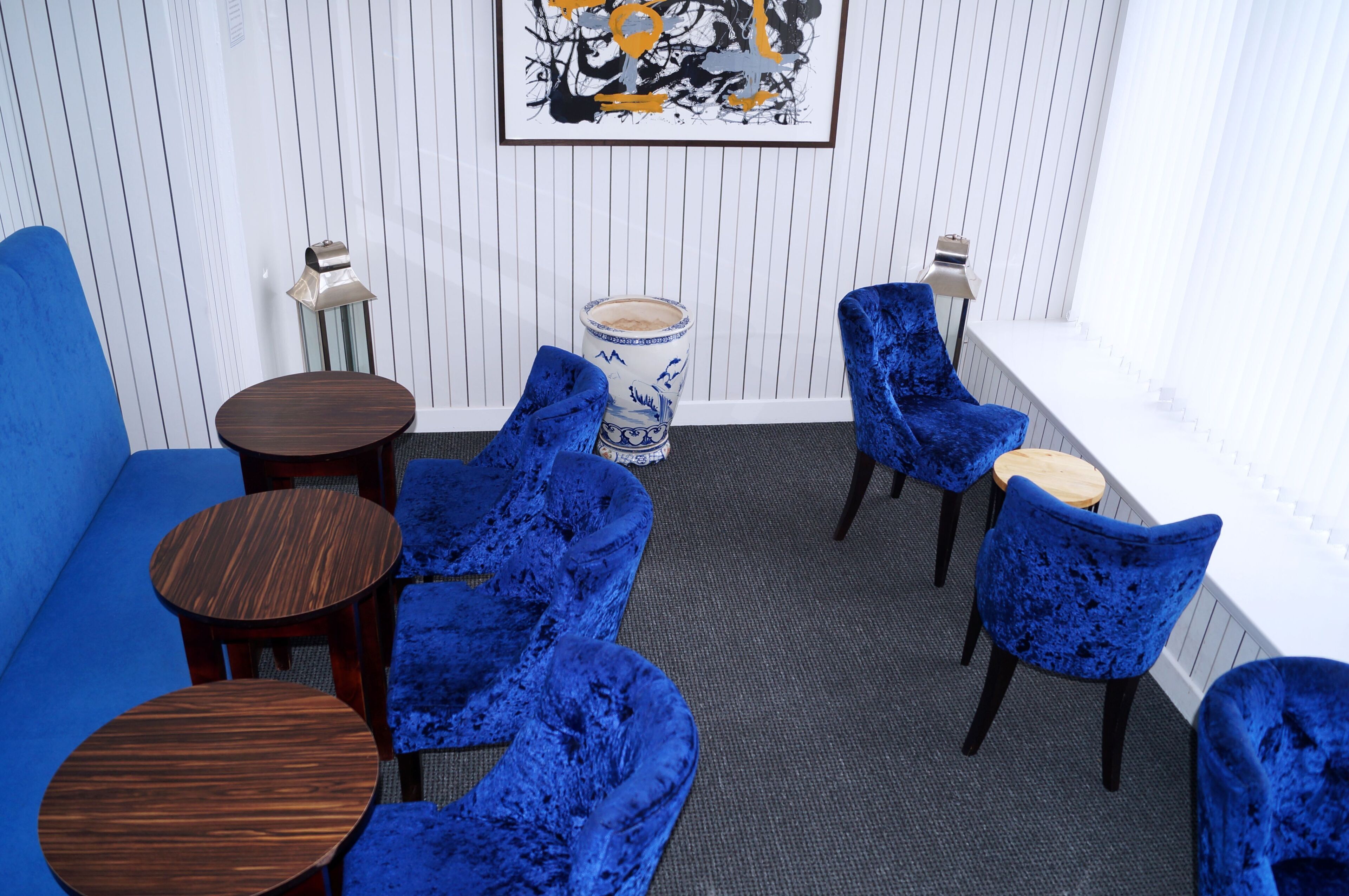 Una habitación con un sofá azul, sillas de terciopelo azul, una mesita de madera, un jarrón decorativo y una obra de arte enmarcada en la pared.