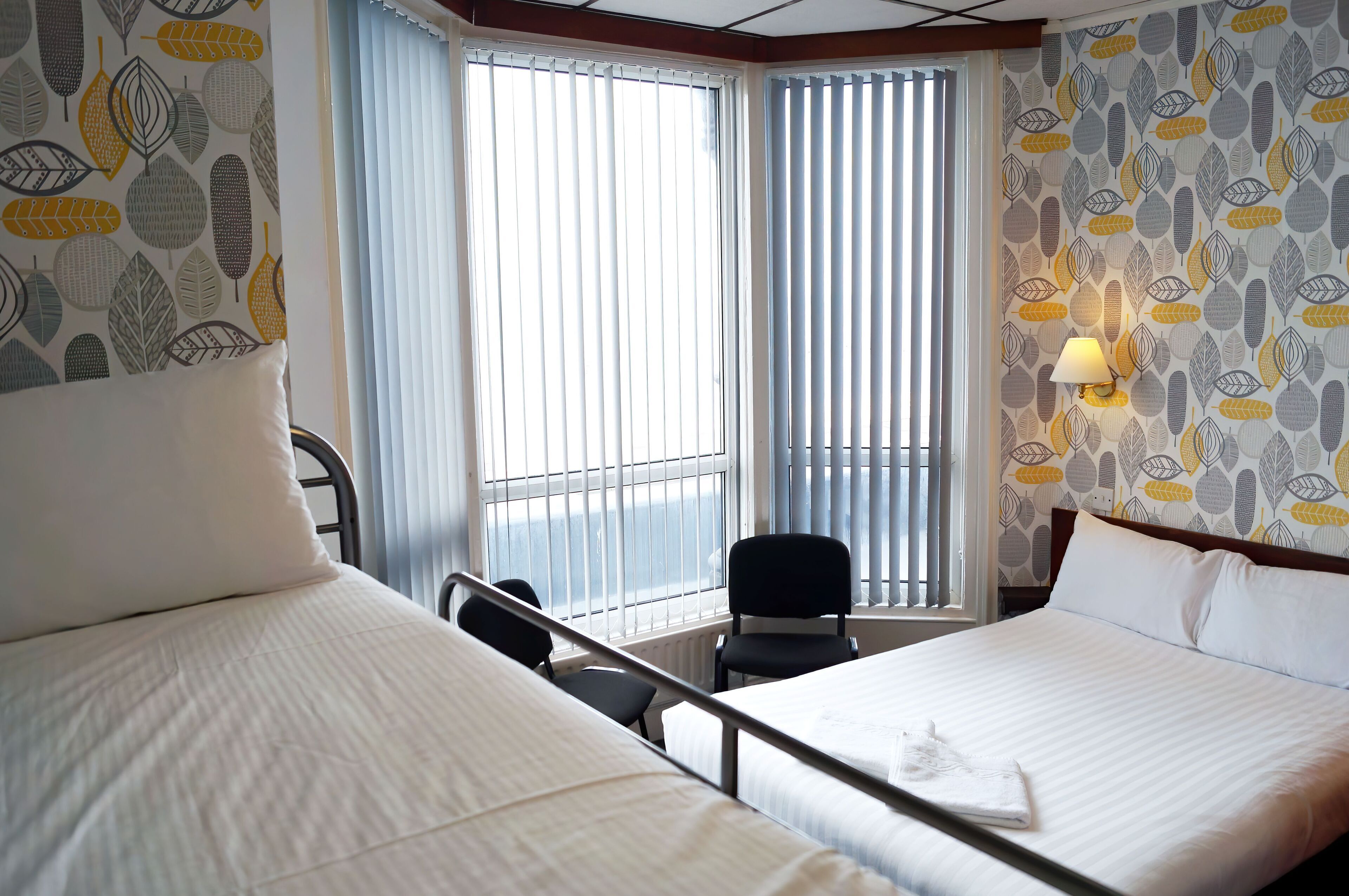 Ein Hotelzimmer mit zwei Betten, einem Sessel, einem Fenster mit Jalousien und einer Wand mit Blättermuster.
