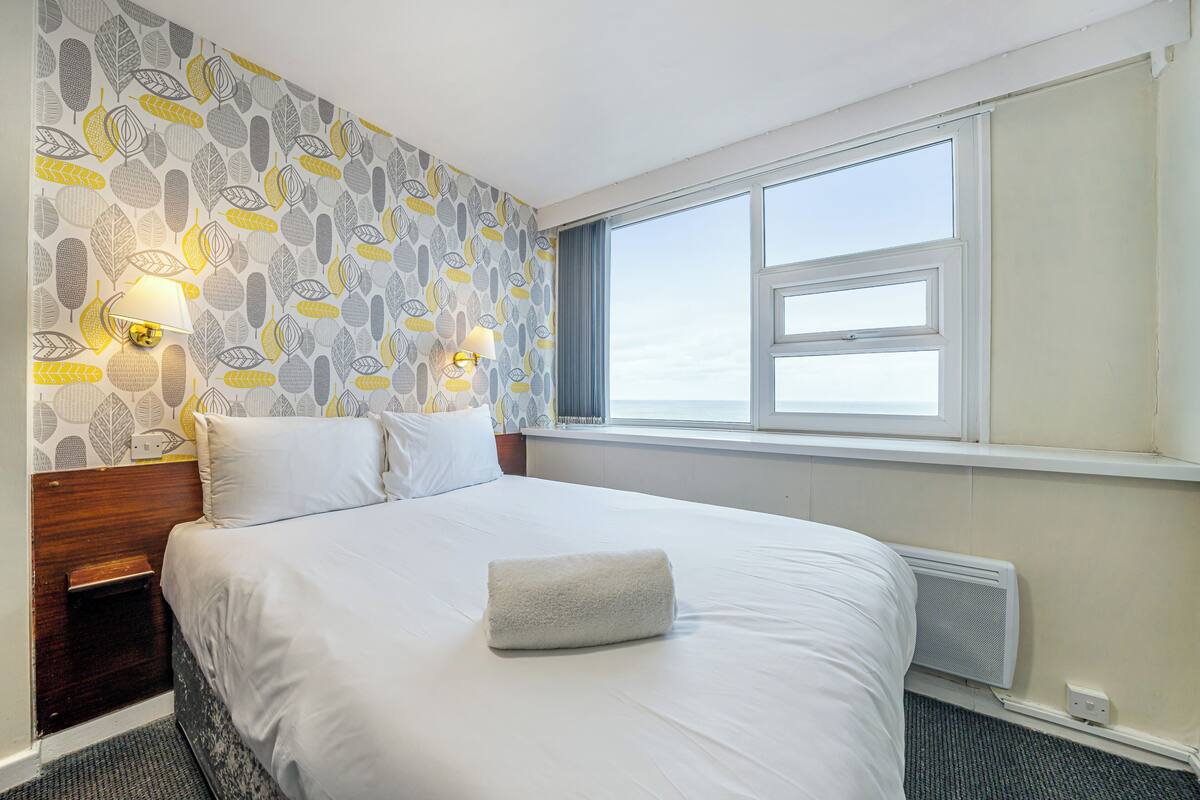 A hotel room with a bed, two bedside lamps, a window with a view, and a patterned wallpaper.