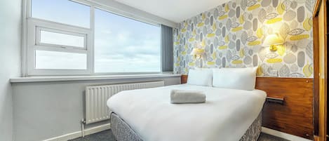 A hotel room with a large bed, a window, a radiator, and patterned wallpaper.