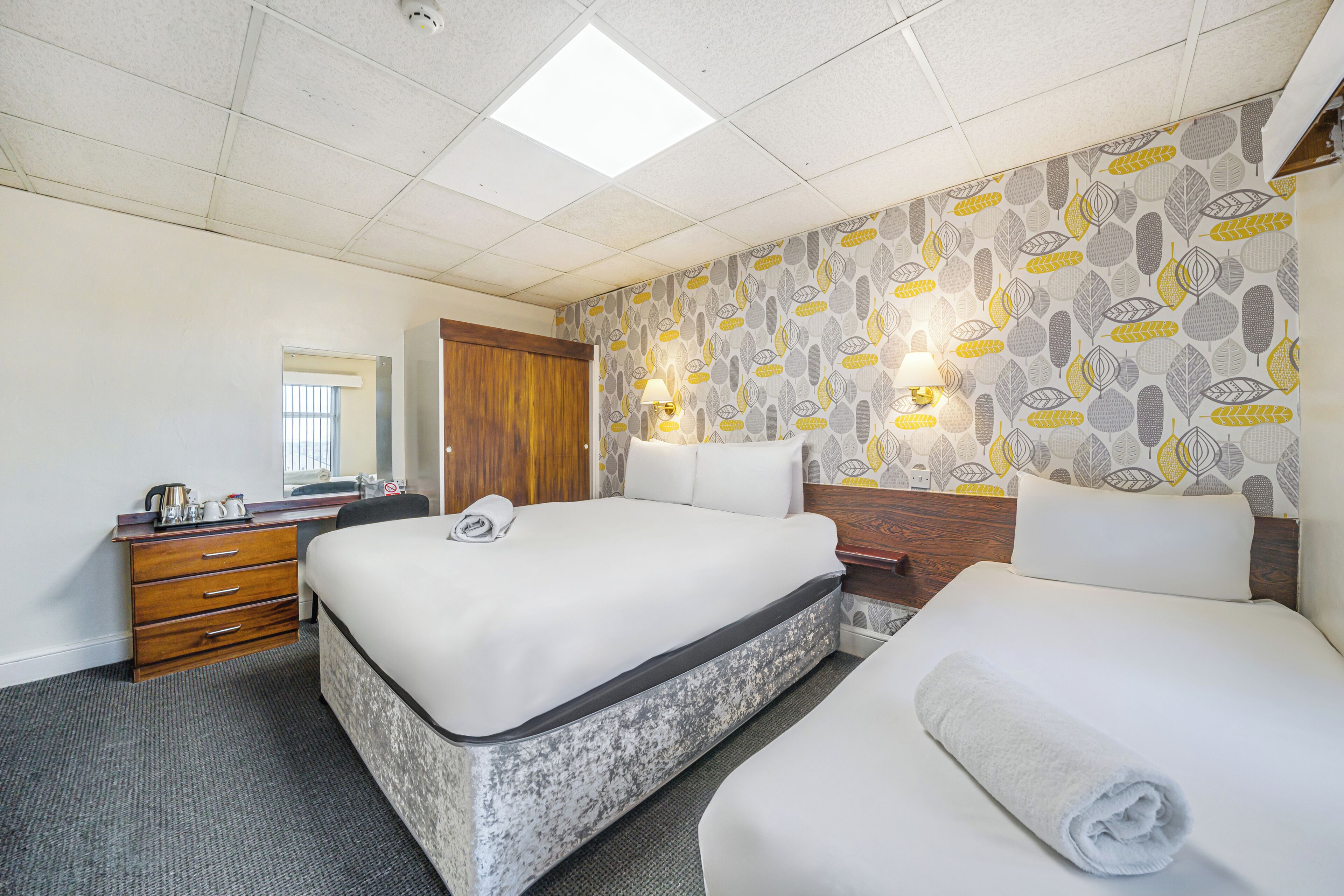 A hotel room with two beds, a wooden dresser, a desk, a mirror, and patterned wallpaper.