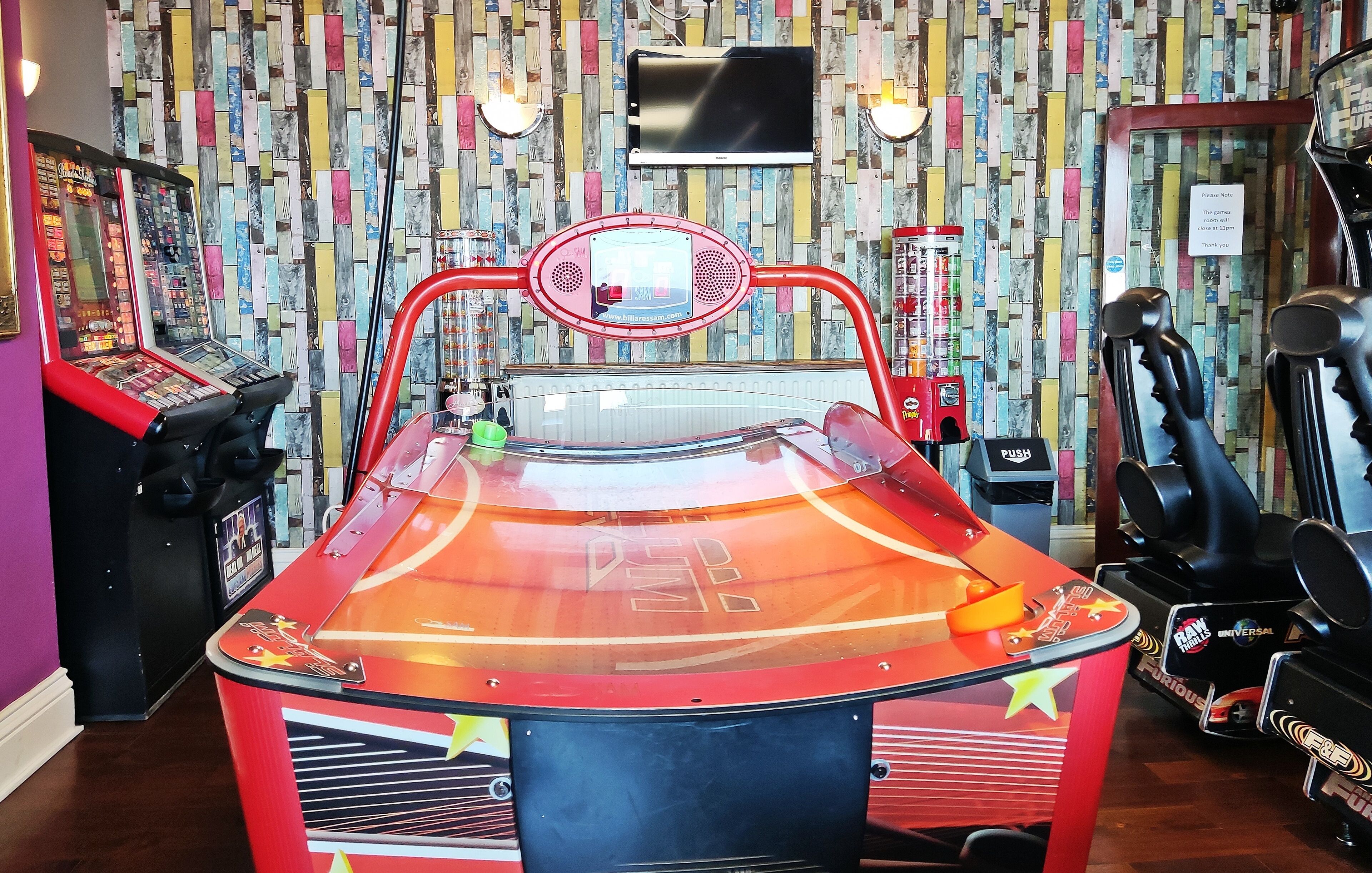 Una máquina de pinball en una habitación con una pared de diseño llamativo y un televisor montado.