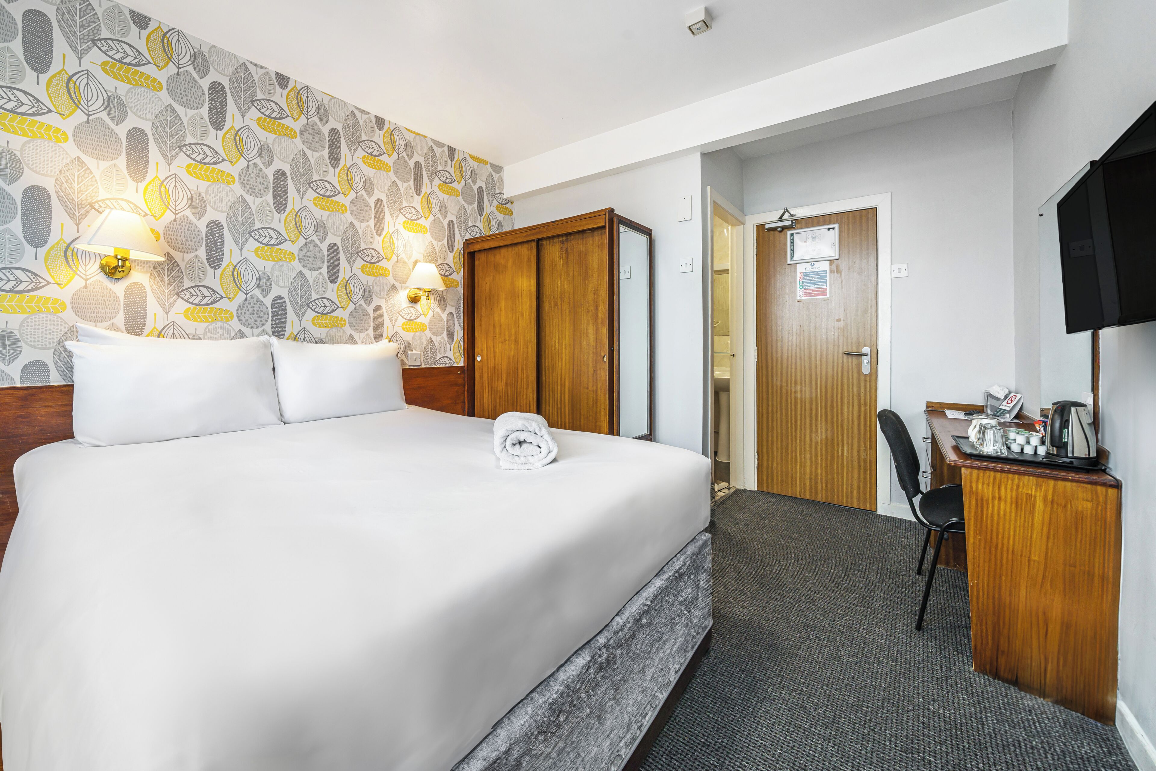 A hotel room with a large bed, a desk with a chair, a TV, and a patterned wallpaper.