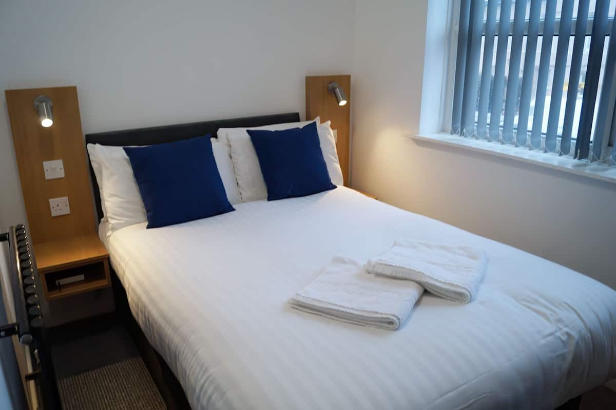 A neatly made bed with white and blue bedding, a bedside table with a lamp, and a window with blinds.