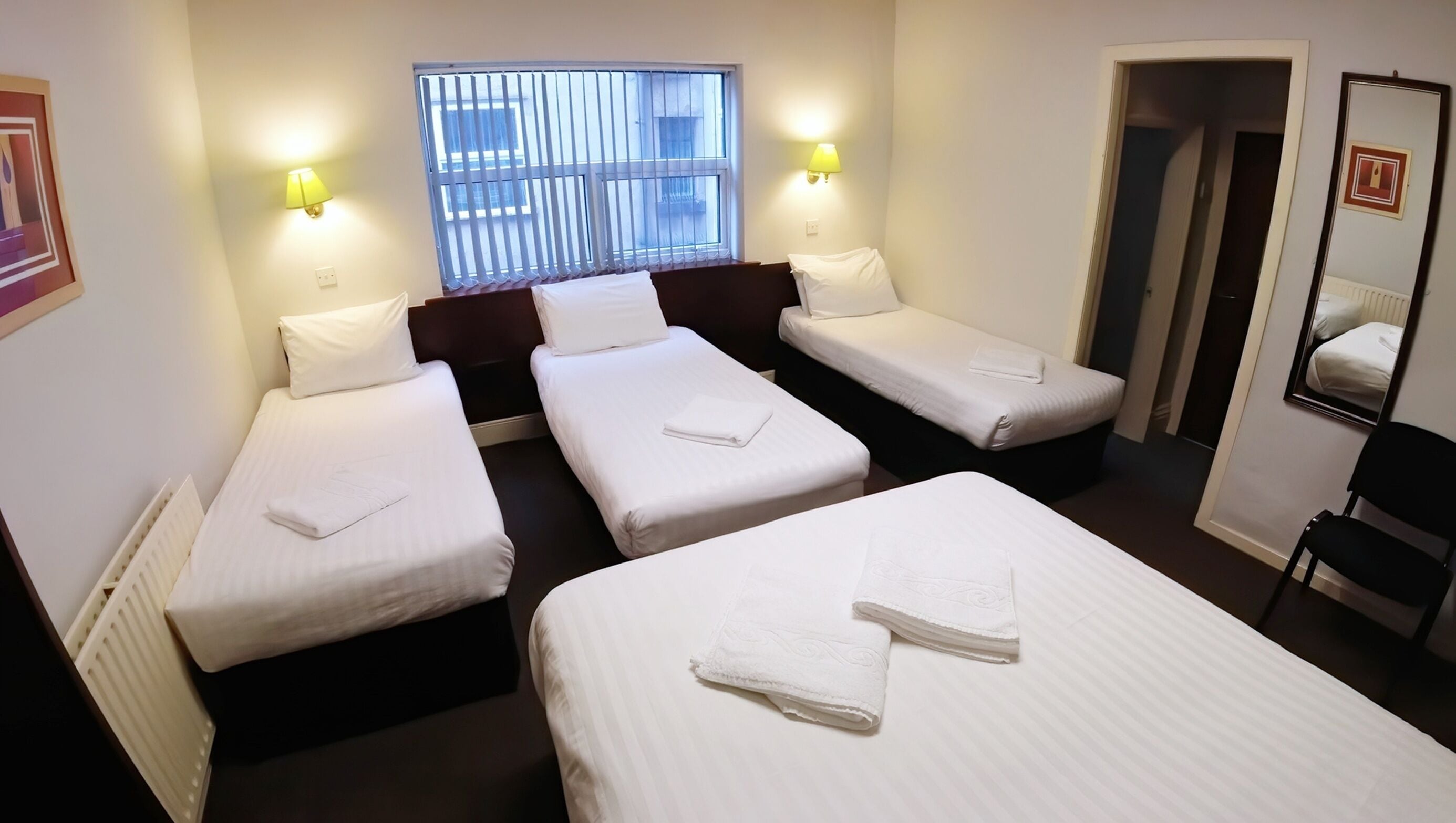 Hotelkamer met drie eenpersoonsbedden, een raam met zonwering, twee wandlampen en een deur naar een andere kamer.