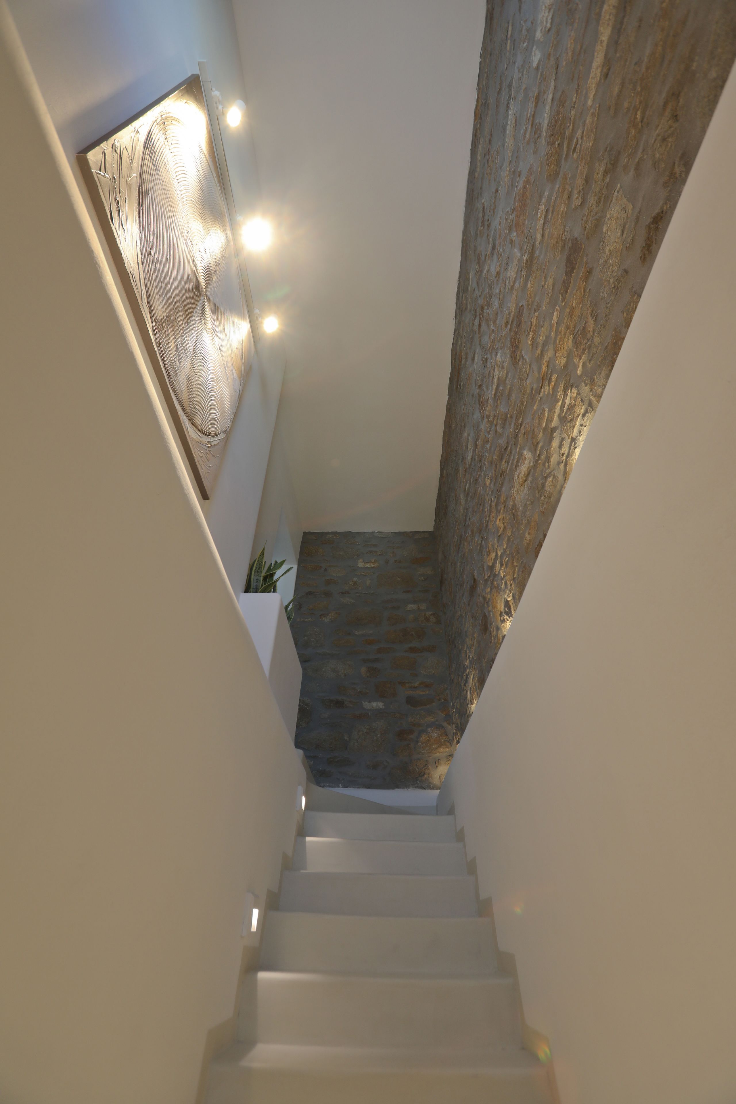Un escalier étroit, avec un mur de pierre d’un côté et un mur blanc de l’autre, mène à un palier doté d’un mur à texture et d’un éclairage encastré.