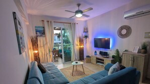 A living room with a blue sofa, a TV, and a ceiling fan. - Andros Apt. 2 Bed, free Wi-Fi, safe, Complex pool (Paralimni)