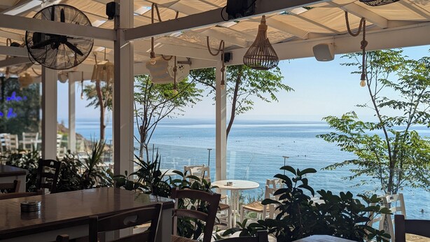 An outdoor dining area with tables and chairs, a view of the sea, and hanging light fixtures. - Andros Apt. 2 Bed, free Wi-Fi, safe, Complex pool (Paralimni)