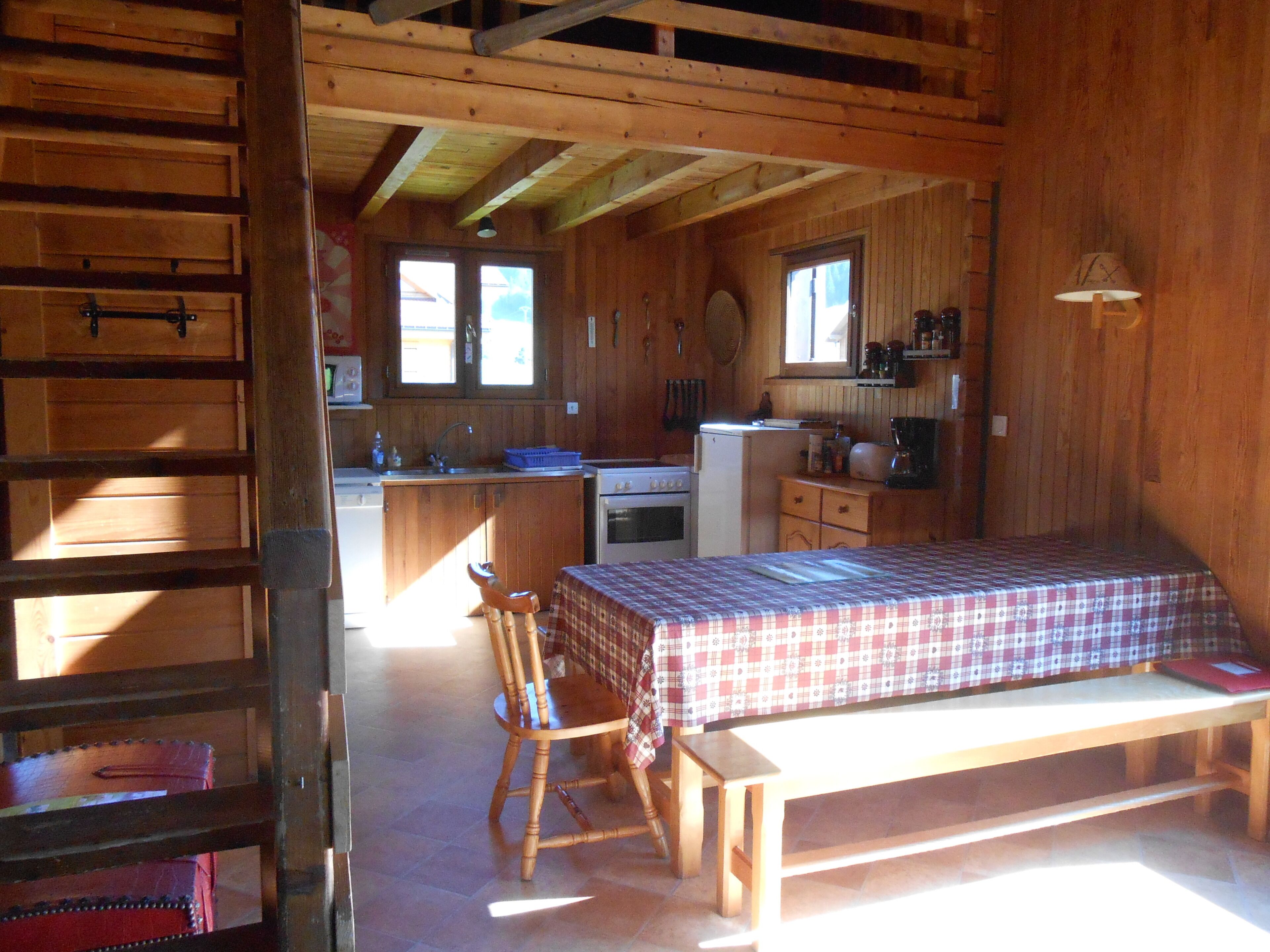 Una habitación compacta de estilo cabaña con interiores de madera, una cama con colcha de cuadros, un banco de madera, una silla, una kitchenette pequeña y una ventana que proporciona luz natural.
