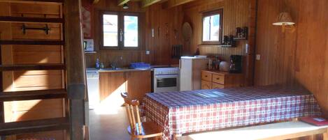 Una habitación compacta de estilo cabaña con interiores de madera, una cama con colcha de cuadros, un banco de madera, una silla, una kitchenette pequeña y una ventana que proporciona luz natural.