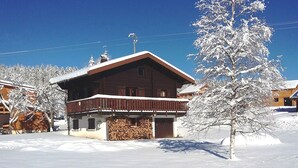 Un chalet en bois doté d’un balcon, entouré d’arbres couverts de neige et surplombant un ciel bleu clair.