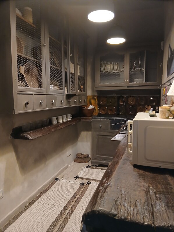 Una cocina con microondas, estufa y diversos utensilios. Hay un estante de madera y una alfombra en el suelo.