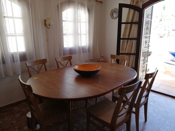 Un coin repas comprenant une table ronde en bois, six chaises en bois, un grand saladier et une porte avec un portail en métal.
