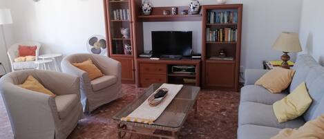 Een woonkamer met een bank, twee fauteuils, een salontafel, een televisie en een boekenkast.