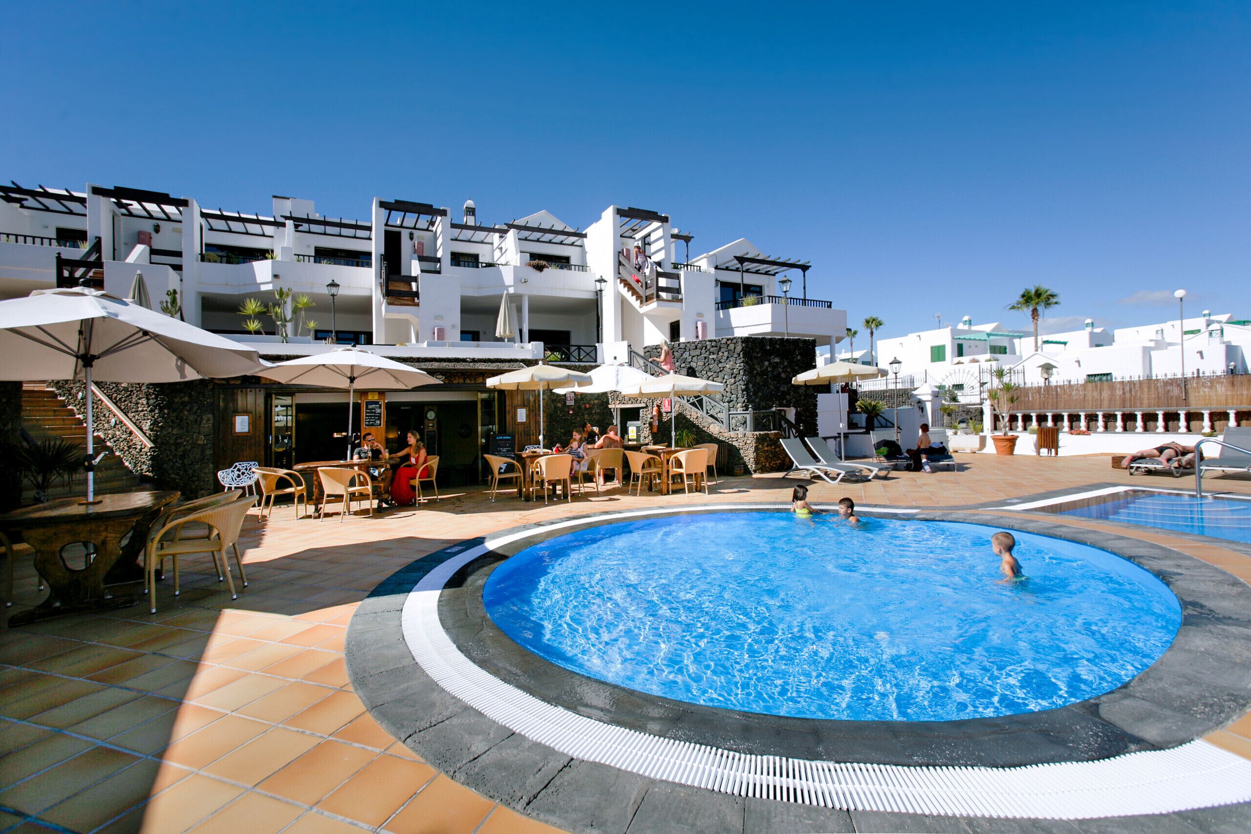 A swimming pool area with lounge chairs and umbrellas, surrounded by a building with multiple balconies and outdoor seating.
