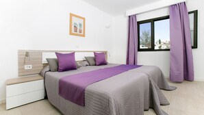 A bedroom with two beds, a window with purple curtains, a bedside table, and a wall-mounted light. - Club Oceano 1 bedroom apts. (Puerto del Carmen)