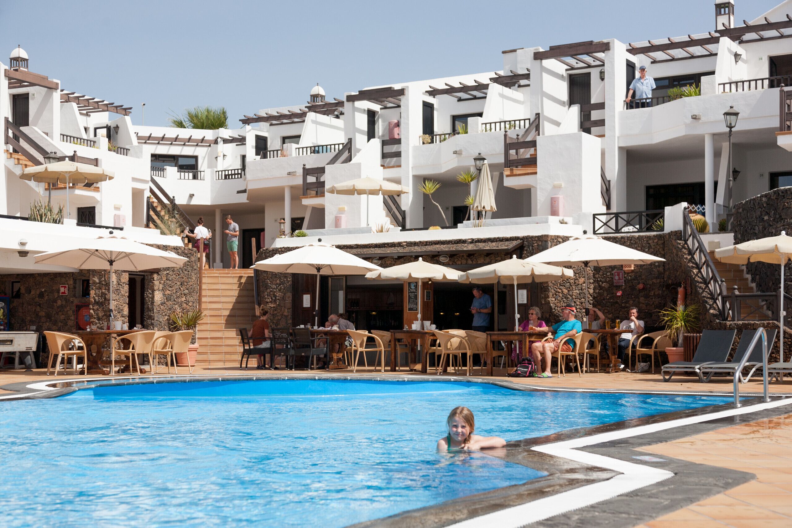A swimming pool area with lounge chairs and umbrellas, surrounded by white buildings with balconies.