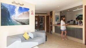 A reception area with a large wave painting, a reception desk, a grey sofa with cushions, and a tiled floor. - Club Oceano 1 bedroom apts. (Puerto del Carmen)