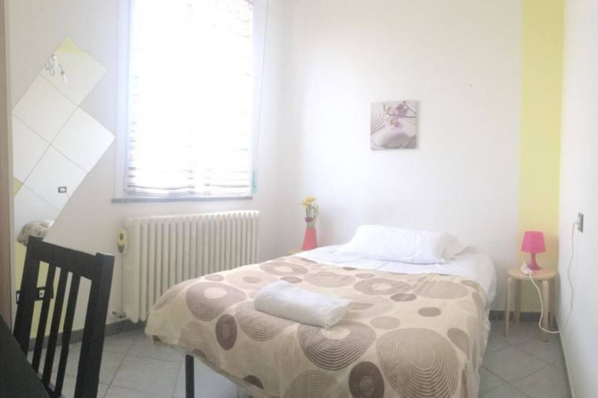 A bedroom with a bed, a chair, a small table, a radiator, and a window.