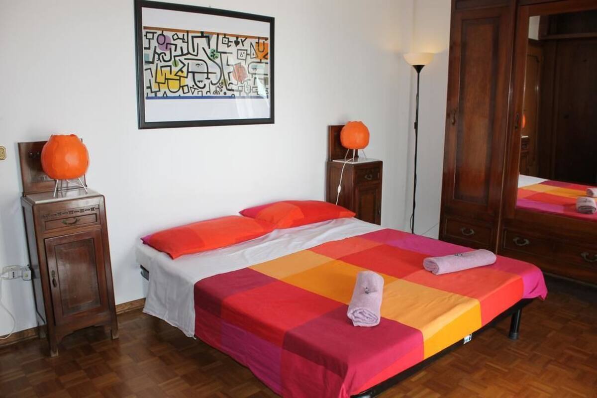 A hotel room with a bed, a bedside table, a lamp, a wardrobe, and a framed artwork on the wall.