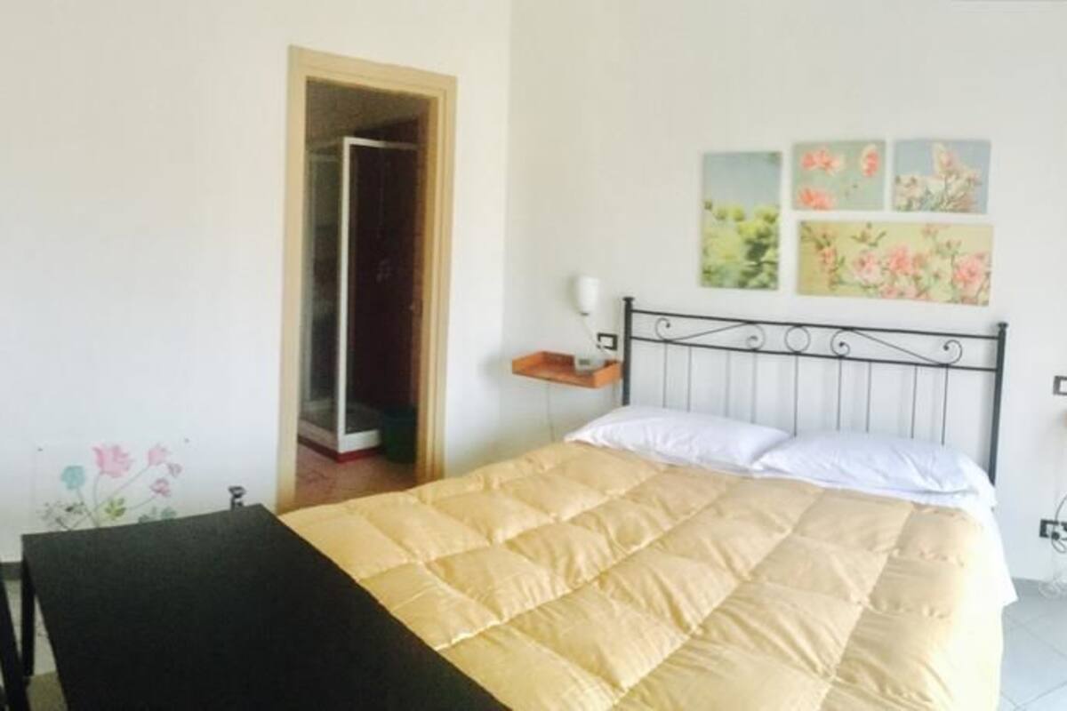 A bedroom with a bed, a wooden door, a chair, a small table, and a wall-mounted shelf.