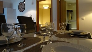Zona de menjador amb una taula preparada per a quatre, incloent copes de vi, plats i cubertries. Hi ha un rellotge a la paret i una porta que dona a una altra habitació.