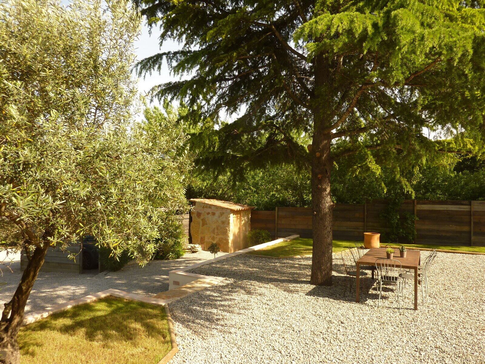 Un patio de grava con una mesa y sillas de madera, rodeado de árboles y una caseta pequeña.