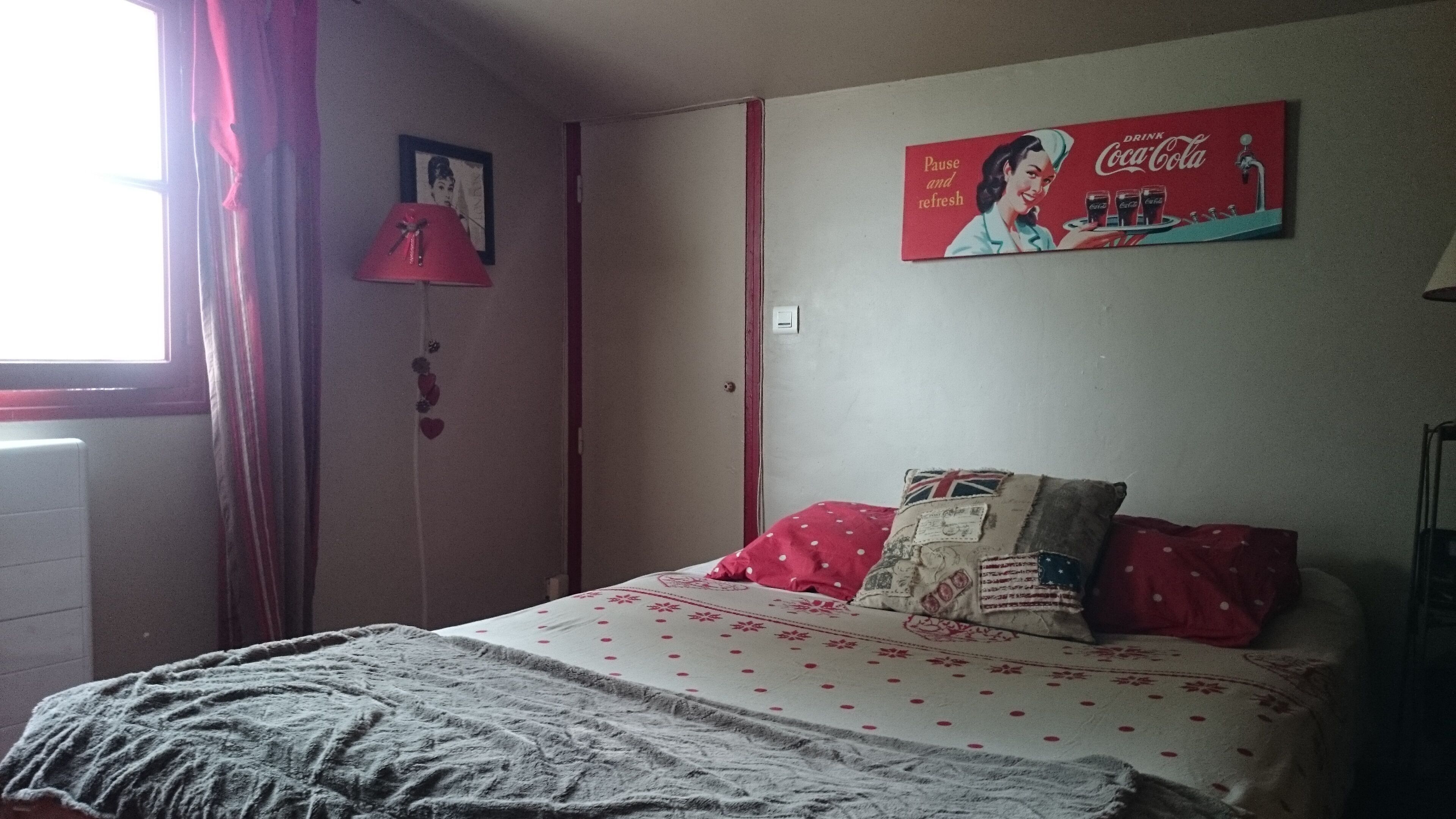 A bedroom with a bed, a window with curtains, a wall-mounted lamp, a framed picture, and a Coca-Cola advertisement.