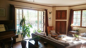 A living room with a sofa, armchairs, a coffee table, and a piano. There are potted plants and a bookshelf with books.