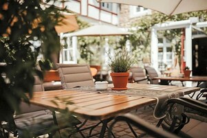 Un patio al aire libre con mesas y sillas de madera, plantas en macetas y sombrillas.