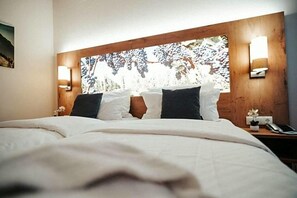 A hotel room with a large bed, two wall-mounted lights, a wooden headboard with a nature-themed artwork, and a nightstand with a phone and a small plant. - Rodderhof (Bad Neuenahr-Ahrweiler)