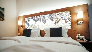 A hotel room with a large bed, two wall-mounted lights, a wooden headboard with a nature-themed artwork, and a nightstand with a phone and a small plant. - Rodderhof (Bad Neuenahr-Ahrweiler)