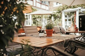 An outdoor patio with wooden tables and chairs, potted plants, and umbrellas.