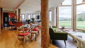 Ein großzügiger Wohnbereich mit Esstisch, Stühlen und einem grünen Sofa. In der Mitte befindet sich eine große Holzsäule, im Hintergrund ist eine Küchenzeile erkennbar.