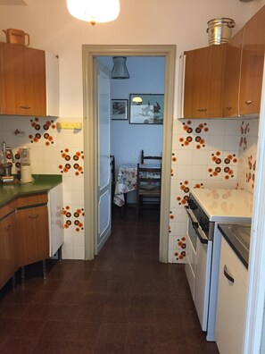 Compact kitchen with sink, stove, and countertop, featuring a kettle and a doorway to a cozy bedroom with a bed and nightstand.