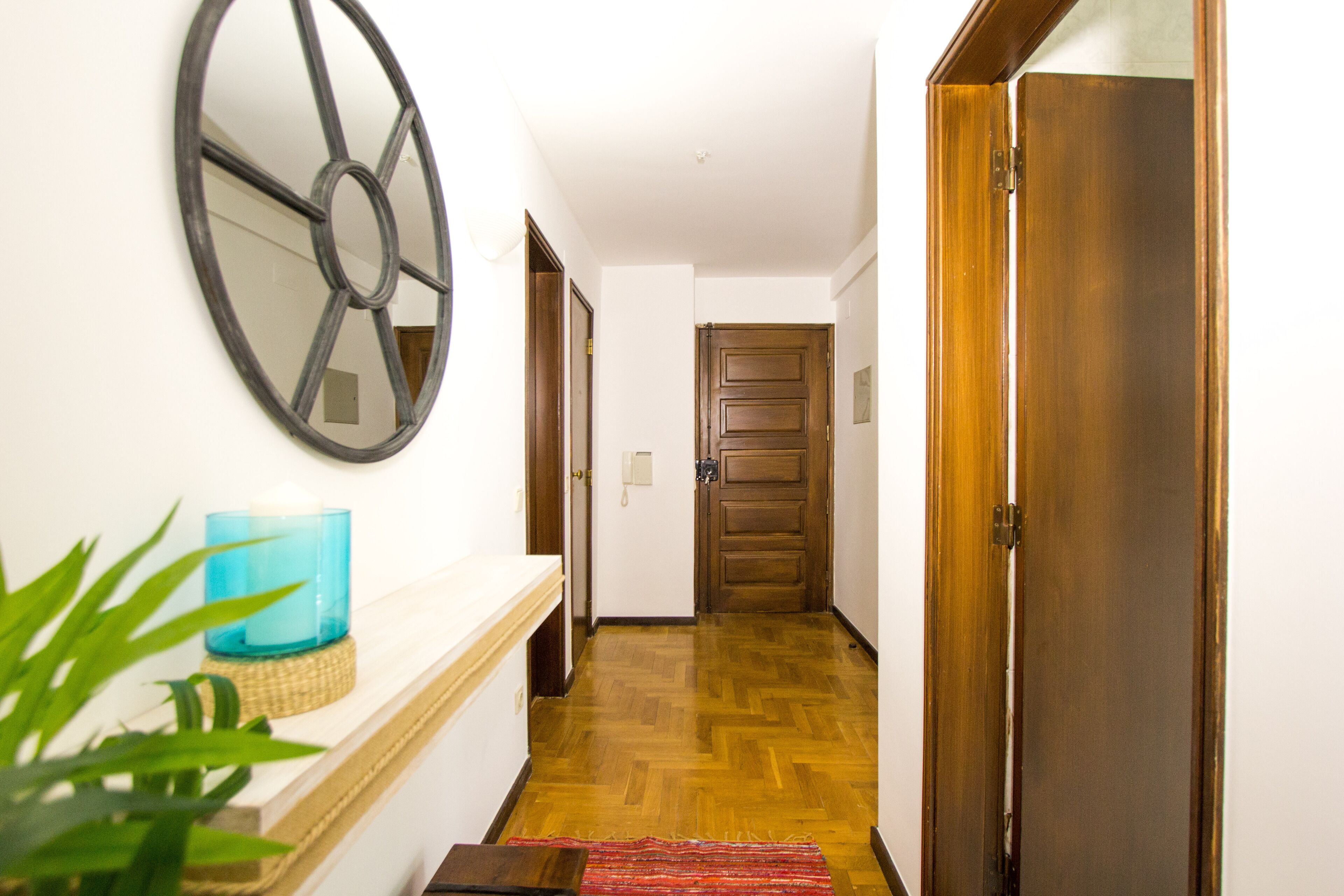 Un pasillo con piso de madera, un espejo, un objeto de vidrio azul sobre una estantería y una puerta al final.